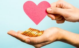 Hand showing a red heart paper shape and the other hand holding a pile of coins, all on blue background.