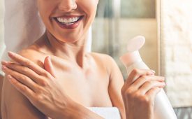 Cropped image of beautiful young woman in bath towel applying body lotion on her shoulders and smiling while standing in bathroom