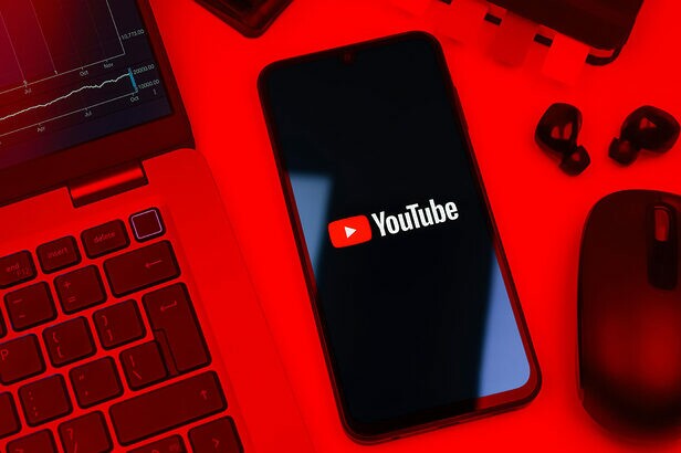 Smartphone com logo do YouTube, laptop e mouse em ambiente vermelho.