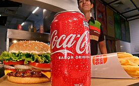 Lata de Coca-Cola gelada, hambúrguer e batatas fritas em balcão de fast-food com funcionário ao fundo.