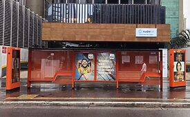 Abrigo de ônibus urbano com painéis publicitários e pessoa.