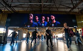 Telão digital exibe jogadores de futebol em ambiente público com pessoas caminhando.