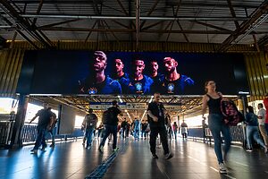 Telão digital exibe jogadores de futebol em ambiente público com pessoas caminhando.
