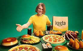 Homem sorridente com mesa cheia de comida variada e sacola Keeta em fundo verde.