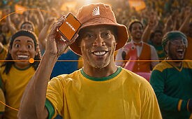Ex-jogador de futebol Ronaldinho Gaúcho com chapéu e uniforme amarelo, segurando um pequeno aparelho eletrônico.