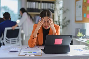 Mulher com blazer laranja, estressada, mãos na cabeça, tablet e papéis na mesa.
