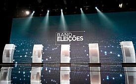 Cenário de debate político na Band, com púlpitos iluminados e logo Band Eleições ao fundo.