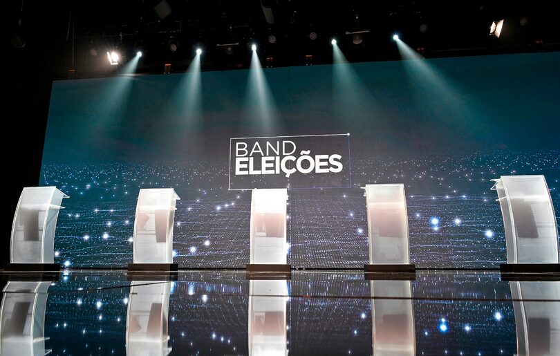 Cenário de debate político na Band, com púlpitos iluminados e logo Band Eleições ao fundo.
