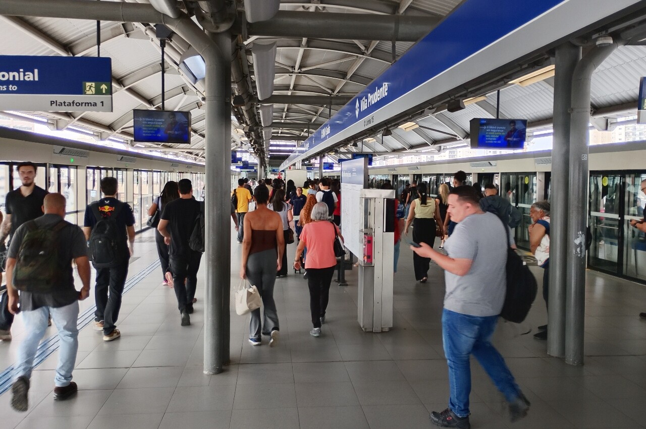 Vista da plataforma de metrô com passageiros caminhando e esperando, portas do trem fechadas.