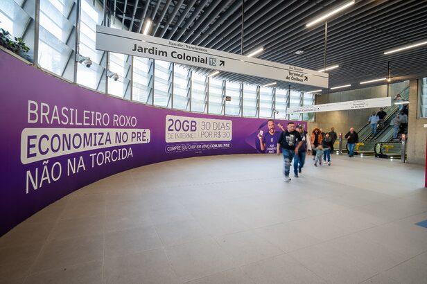 Estação de metrô moderna com letreiro "Jardim Colonial" e painel roxo de publicidade de celular com pessoas.
