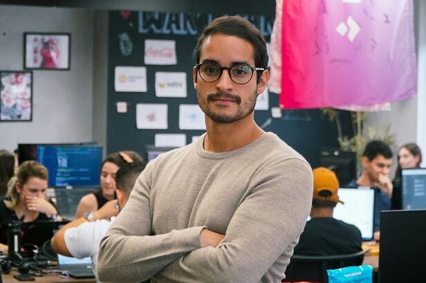 Homem com óculos e camisa bege, braços cruzados, em escritório com computadores e painel de ideias.