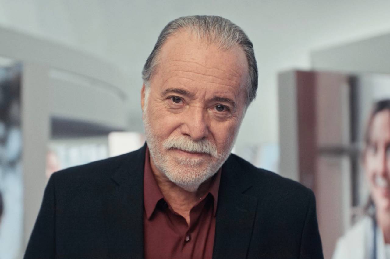 Homem idoso com cabelo e barba grisalhos, camisa vinho e blazer preto, olhando para frente.