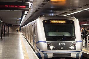 Metrô prateado e preto na plataforma de uma estação, com passageiros esperando e detalhes como piso tátil.