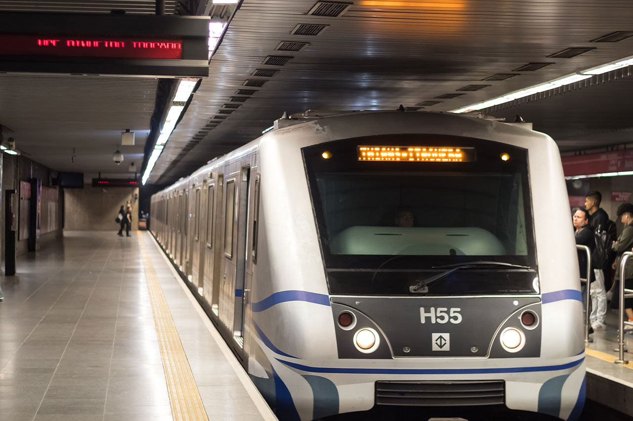 Metrô prateado e preto na plataforma de uma estação, com passageiros esperando e detalhes como piso tátil.