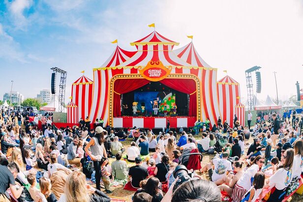 Festivalzinho 2025 - Grande tenda de circo vermelha e branca, palco com show de bonecos para público sentado em festival ao ar livre.