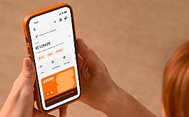 Mãos segurando smartphone laranja exibindo aplicativo do Banco Inter com saldo de R$ 1.234,00.