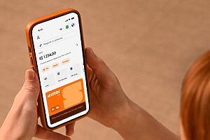 Mãos segurando smartphone laranja exibindo aplicativo do Banco Inter com saldo de R$ 1.234,00.
