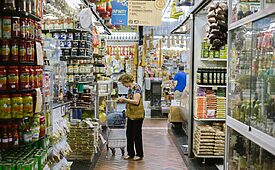 Mulher em mercado com carrinho, olhando produtos em prateleiras cheias de alimentos.