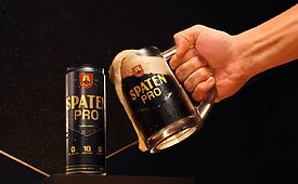 Mão segurando caneca Spaten Pro transbordando cerveja escura e espuma; lata de Spaten Pro ao lado.