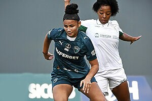 Duas jogadoras de futebol em campo: uma de camisa verde-escura e outra de camisa branca em disputa de bola.