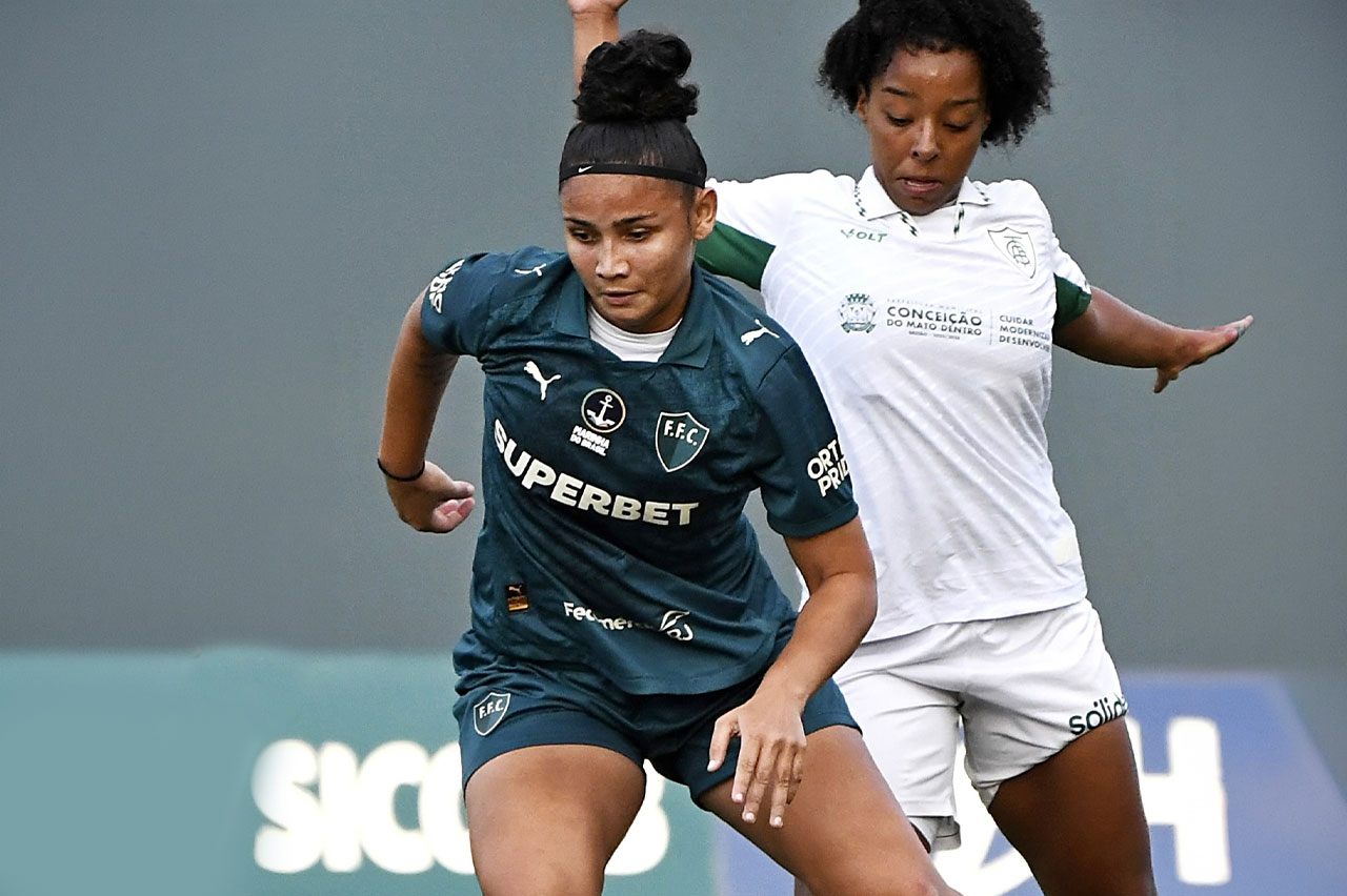Duas jogadoras de futebol em campo: uma de camisa verde-escura e outra de camisa branca em disputa de bola.