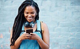 Jovem mulher negra com dreads sorrindo, olhando o celular com fones no pescoço, roupa jeans.
