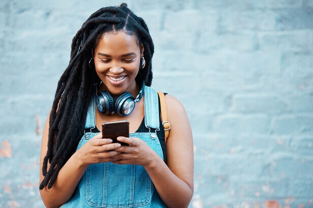 Jovem mulher negra com dreads sorrindo, olhando o celular com fones no pescoço, roupa jeans.