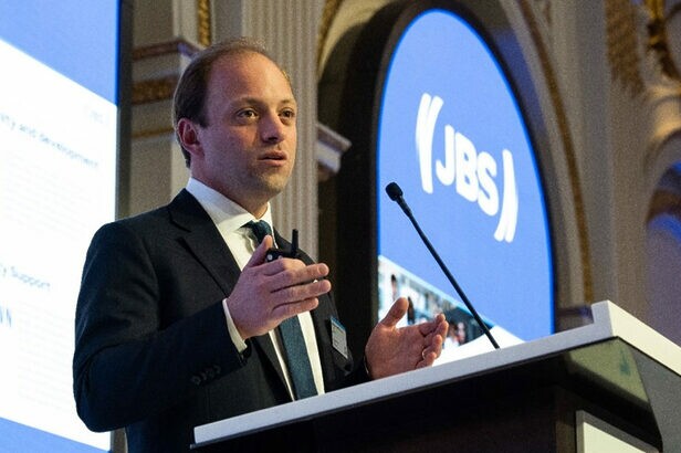 Orador gesticulando em apresentação; logo JBS visível em painel luminoso.