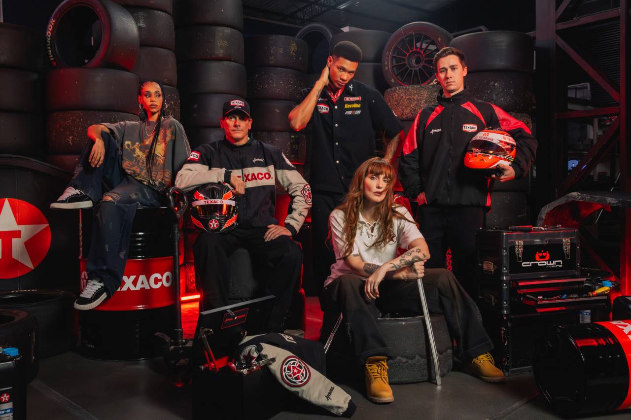 Jovens em uniformes automotivos em cenário de garagem com pneus, capacetes e barris de óleo Texaco.