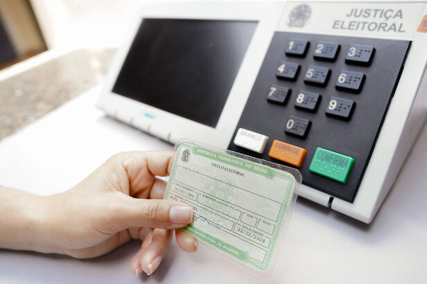 Mão segurando título de eleitor com urna eletrônica da Justiça Eleitoral ao fundo.