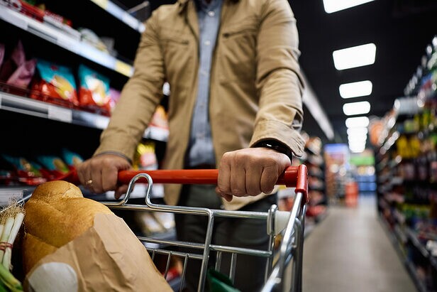 Homem empurrando carrinho de compras com pão e vegetais em supermercado, entre prateleiras de produtos.