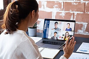Mulher em videoconferência no laptop com quatro participantes, segurando caneta, em mesa com notebook.