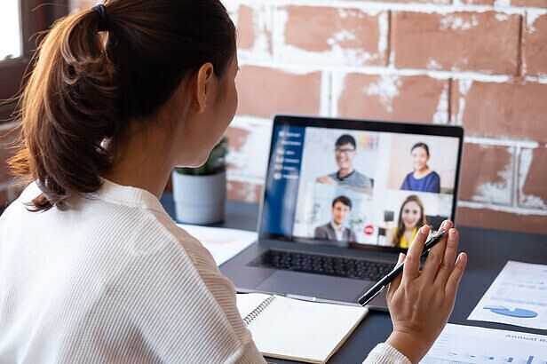 Mulher em videoconferência no laptop com quatro participantes, segurando caneta, em mesa com notebook.