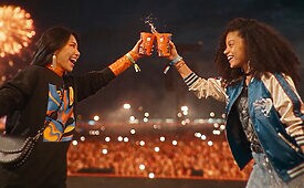Duas amigas brindam com copos laranja em festival noturno, fogos de artifício e público ao fundo.