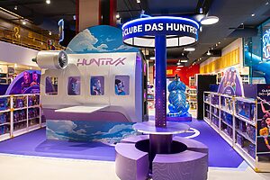 Área temática "Clube das Huntrix" roxa e branca, com réplica de avião, dentro de uma loja.