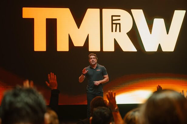 Homem em palco escuro apresentando evento "upfront eletromidia 2027 TMRW" em telão digital grande.