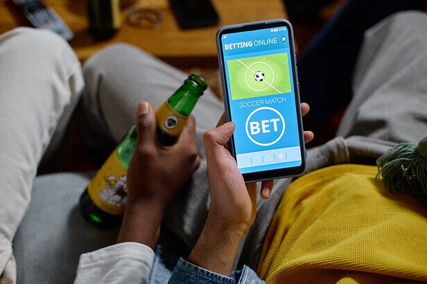 Pessoa segurando smartphone com aplicativo de apostas de futebol e garrafa de cerveja.