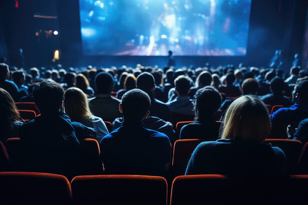 Pessoas assistindo a um filme em um cinema escuro, vista das costas dos espectadores.