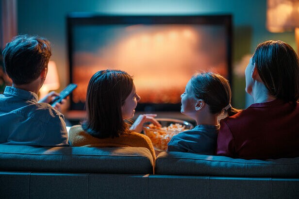 Quatro pessoas juntas no sofá olhando para uma TV, com um balde de pipoca no centro.